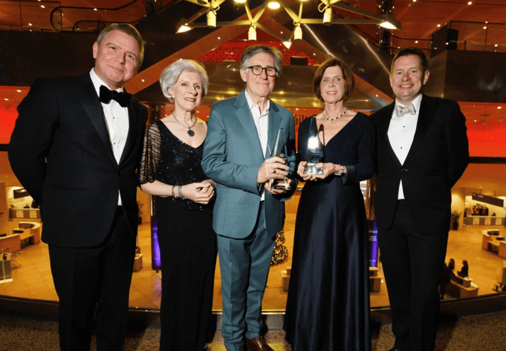 A formal group photograph of five distinguished professionals at the Business & Finance Awards event. From left to right, Ian Hyland, President and Publisher of Business & Finance in a black tuxedo and bow tie,Professor Frances Ruane, Chair of The National Competitiveness Council with short gray hair in a black evening gown with sheer sleeves, actor and writer Gabriel Byrne holding an award in a blue suit, Dame Louise Richardson, President of the Carnegie Corporation New York in a navy blue evening gown holding an award, and Seamus Hand, Managing Partner, KPMG Ireland in a black tuxedo and bow tie. They are all smiling and looking at the camera, with a modern, softly lit venue in the background.