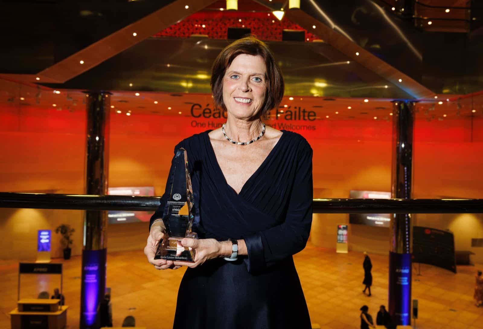 Dame Louise Richardson, dressed in an elegant navy blue evening gown, is holding a clear glass award with a smile. Sutherland Leadership Award