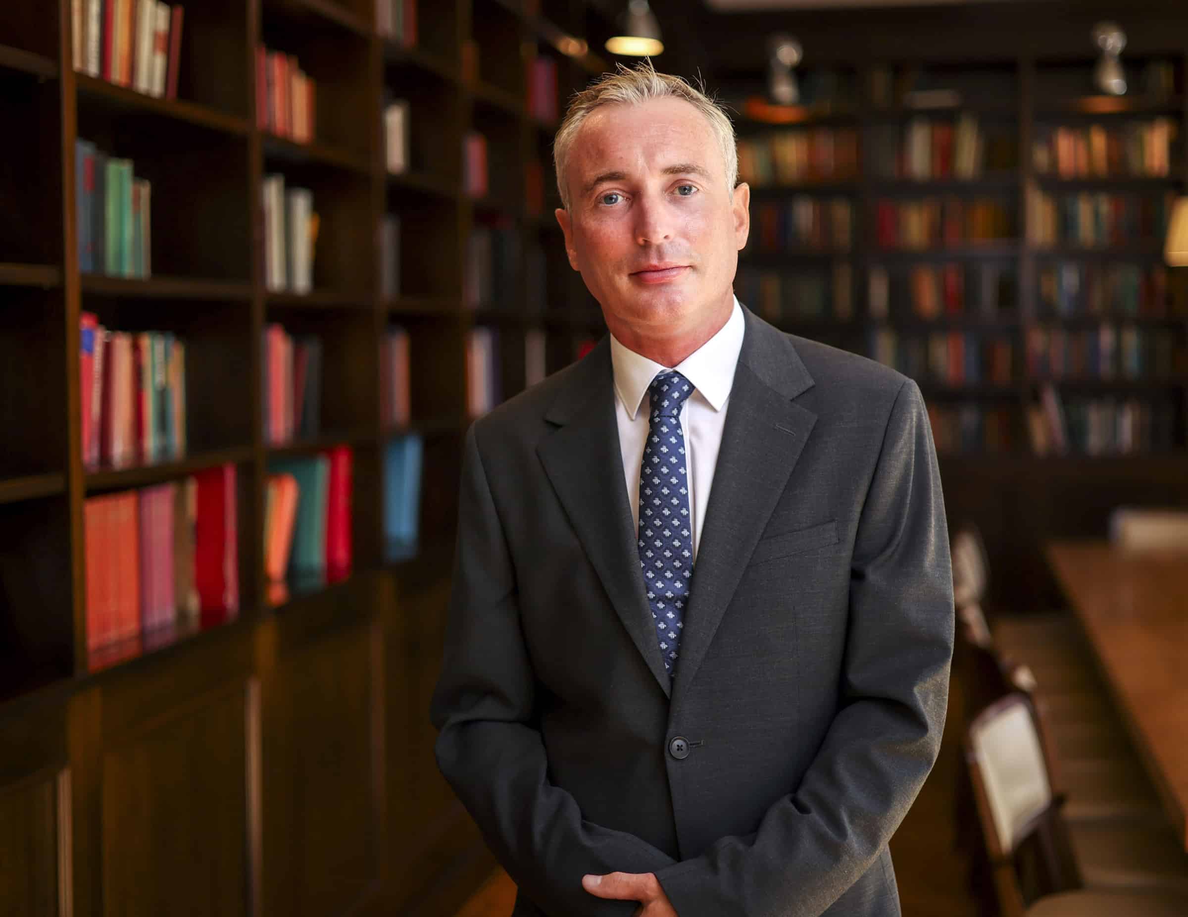 Ian Brady, Chief Executive Officer of Unio Group, standing in a professional setting with a background of bookshelves filled with colorful books. He is dressed in a dark grey suit with a white shirt and blue patterned tie, looking directly at the camera with a confident and composed expression, symbolizing his leadership and expertise in the financial services industry.