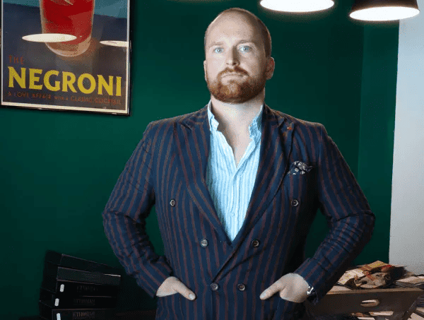Seán Treacy, founder and owner of Hunter Treacy Tailors, standing confidently in front of a green wall, under a vintage 'The Negroni' poster. He is dressed in a finely tailored navy blue pinstripe double-breasted suit, with a light blue unbuttoned shirt