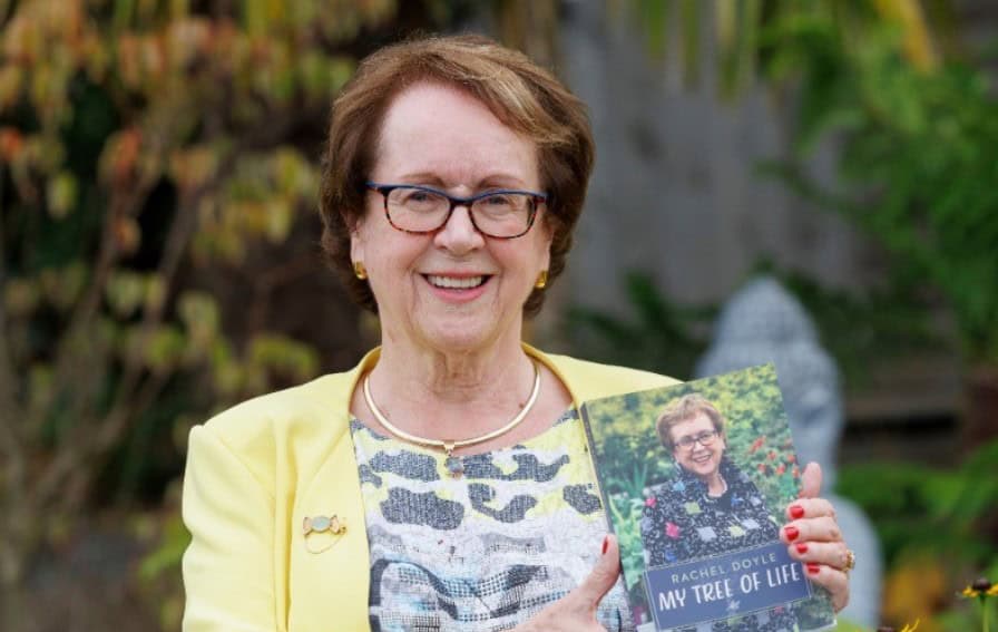 Rachel Doyle, in a yellow blazer and patterned top, smiling as she holds her book 'My Tree of Life' in a garden setting